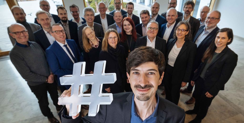 Bei Klick des Bühnensliders kommen Sie auf die Websites der Cluster Offensive. Auf dem Bild sind Mitglieder der Cluster-Offensive zu sehen, die zusammenstehen. Eine Person hält eine Raute hoch, das Symbol der Cluster Offensive.