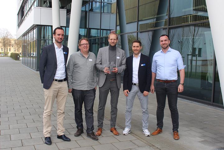 Fünf Männer stehen in Business-Kleidung vor einem modernen Bürogebäude. Der Mann in der Mitte (Sven Blanck, Clustergeschäftsführer) hält eine Trophäe in Form eines Hashtags - das Symbol für die Cluster. Alle lächeln in die Kamera, es handelt sich um ein Gruppenfoto.
