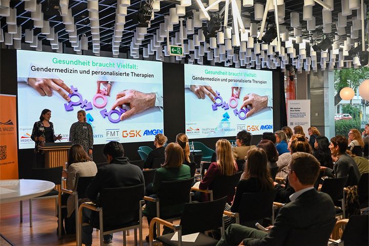 Mehrere Menschen sitzen vor einer Bühne bei der Veranstaltung „Gesundheit braucht Vielfalt“.