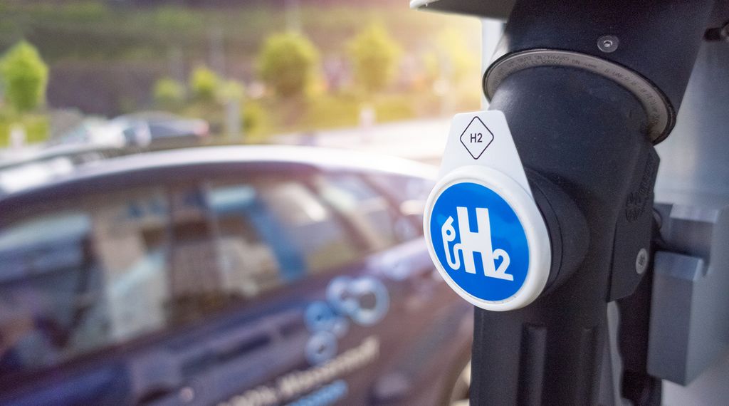 Nahaufnahme einer Wasserstoff-Zapfsäule mit blauem H2-Symbol, im Hintergrund ein parkendes Auto.